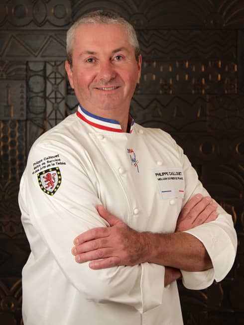 Philippe Caillouet stands with his arms crossed and wearing a white chef's jacket with the colors of the French flag on the collar