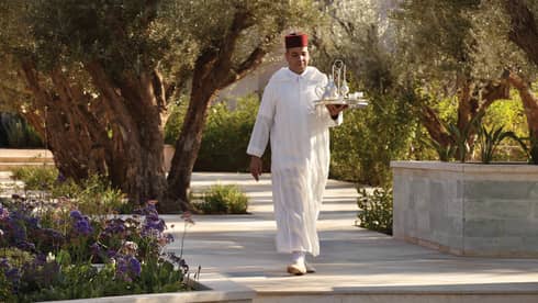 Server carries tray with silver teapots down sunny pathway