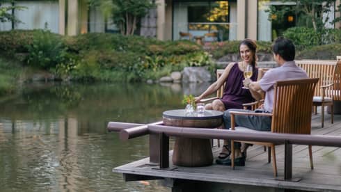 Smiling couple clink champagne flutes at a café table on a wood deck overlooking a rippling pond bordered by lush foliage.