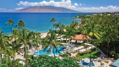 Aerial view of resort pool, patios and palm trees on ocean, mountain in background