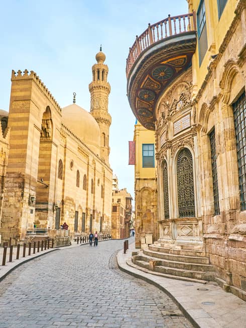 Pedestrians walk along a meandering cobbled road between tall, yellow stone buildings with ornate lattice windows.