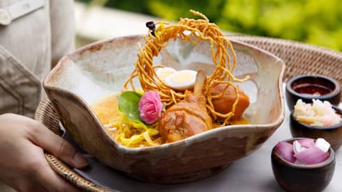 Bowl of curry noodle soup with chicken drumsticks, crispy noodles, boiled egg and garnish, served with condiments on the side.