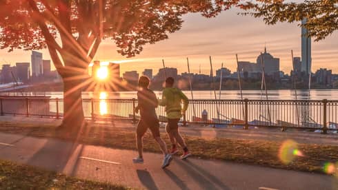 Two people jogging along a tree-lined, riverside walkway with the sun setting behind the Boston skyline