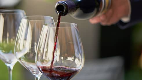 A close up of hands pouring a bottle of red wine into a clear wine glass that rests on a wooden tray, outdoors