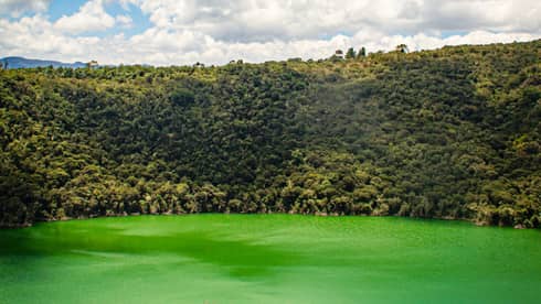 An aerial view of the green-hued waters of Lake Guatavita, surrounded by green trees, with a bright blue cloud-filled sky above
