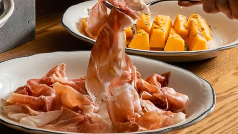 An aperitivo table laden with beautifully plated meats, olives and salads, a fork lifting a slice of prosciutto.