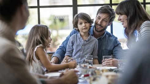 Boy in checkered shirt sits on dad's lap at dining table in sunny room, mom and sister on each side