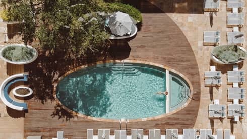 Aerial view of an oval pool surrounded by a rectangular wooden deck and rows of lounge chairs on two sides with greenery and more loungs seating on the other two sides