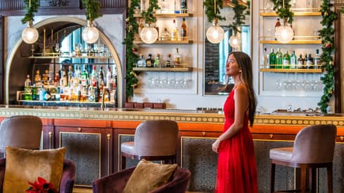A woman in a long red dress stands in the middle of La Capitale restaurant beneath green festive garlands