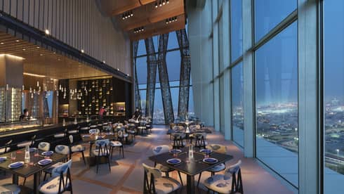 Dimly-lit Sintoho Restaurant at dusk, bar and dining tables and chairs under soaring ceilings, beside glass windows on wall