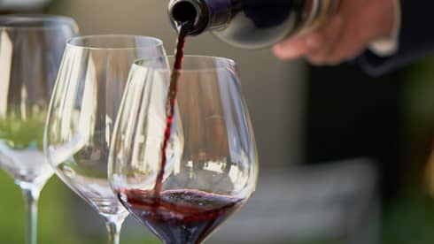 A close up of hands pouring a bottle of red wine into a clear wine glass that rests on a wooden tray, outdoors
