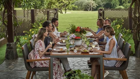 Eight people sit around a long dining table set up outside beneath a canopy of greenery and string lights