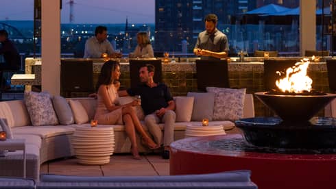 The sky is dark beyond the cinderhouse rooftop bar – furnished with a curved sectional, fire pit, hanging lights and people socialize 
