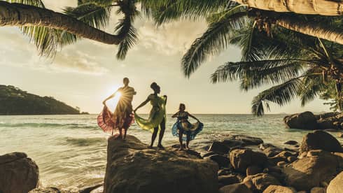 The sun sets over three dancers wearing long, colourful dresses standing on top of rocks by the water