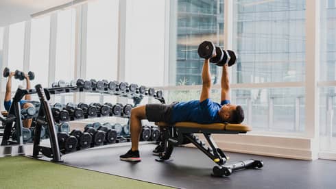 A man working out with free weights in a clean fitness room with large windows.