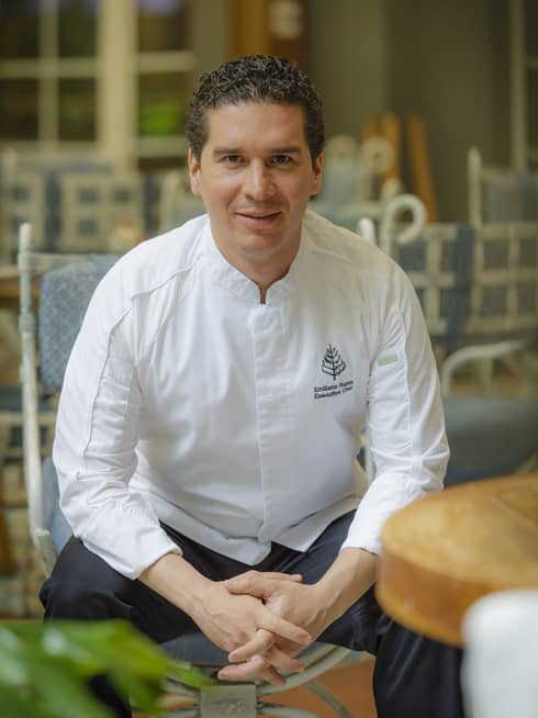 Chef Emiliano Rabia sits on a blue chair resting with his elbows on his knees and hands clasped, staring directly at the camera