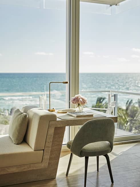 Modern seating area with a table, lamp, and vase of flowers by a large window overlooking a sunny ocean view. A serene and inviting coastal setting.