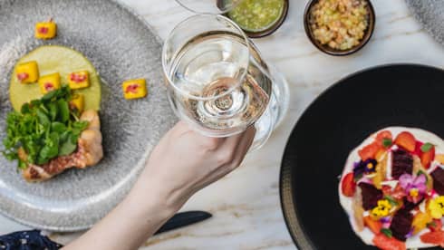 An assortment of food and two people toasting with white wine.