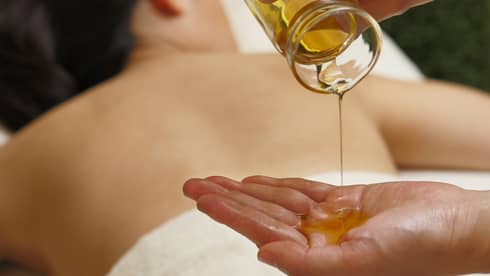 Spa staff pouring massage oil from glass jar in hand over woman lying on table