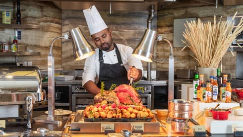 Chefs wearing tall white hat, white shirt and dark blue apron prepares to carve meat at a buffet live station