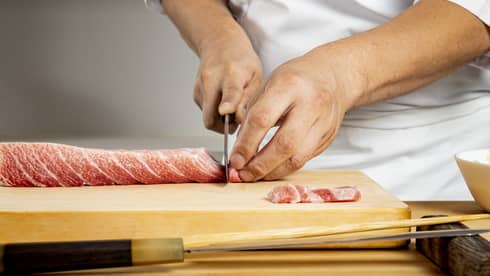 Chef's hands use knife to thinly slice meat on wooden board, preparing it for sushi