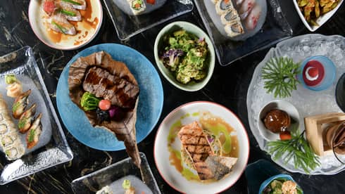 Table filled with various plates of steak, seafood, sushi and desserts presented on glass platters and blue or white dishes