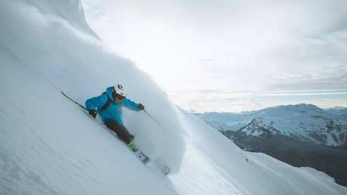 Skier in blue jacket, helmet skis down slope, snow spraying behind him