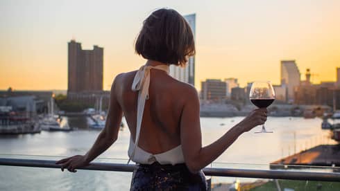 Person with bob haircut wearing a white halter-neck top and dark skirt stands silhouetted at sunset as they hold a glass of wine and overlook a city harbour
