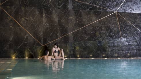Woman sits on edge of indoor swimming pool, man wades beside her 