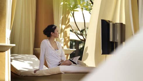 Woman with open book sits on padded patio lounge chair under canopy