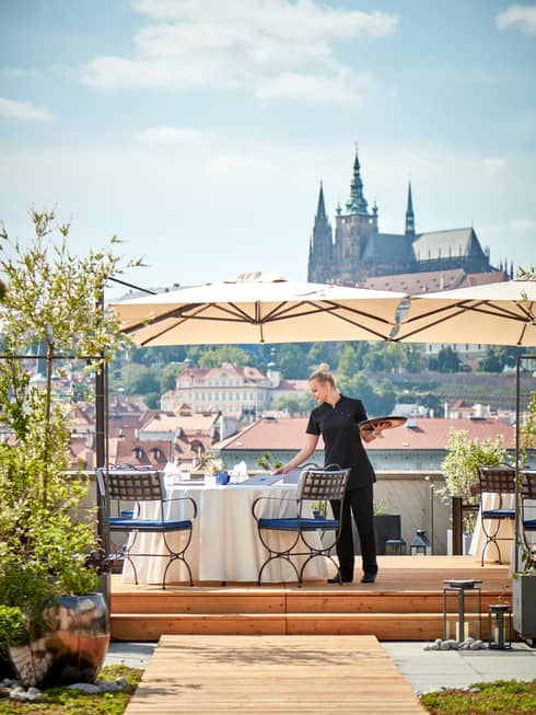 Server with tray in hand beside table with umbrella on rooftop restaurant, Prague Castle in view