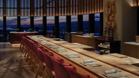 Ten place settings at a long wooden table with warm underlighting, a curved bank of windows offering a dramatic sunset view.