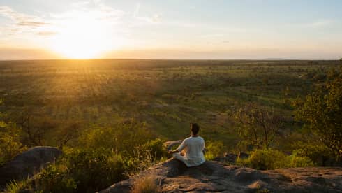 Woman sits crossed-legged, meditates on cliff overlooking Serengeti sunset