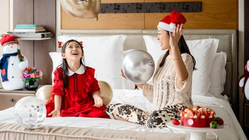 Adult and child adorned in Christmas outfits and hat sit atop bed in guest room with balloons and other holiday décor