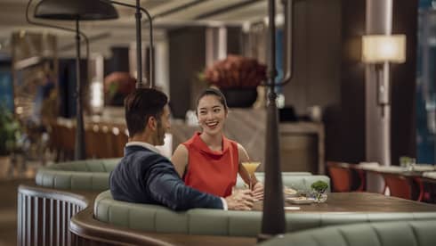 A smiling couple sit at a circular booth in an empty, dimly lit restaurant with cocktails and appetizers on their table.
