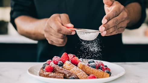 Four slices of French toast topped with raspberries, blueberries and strawberries being sprinkled with icing sugar through small sieve, on white plate on top of white marble counter
