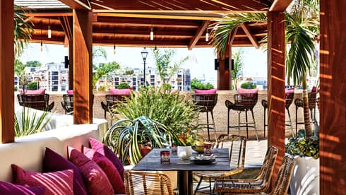 An outdoor eating venue with red cushions on benches and stools surrounded by red pillars and trees.