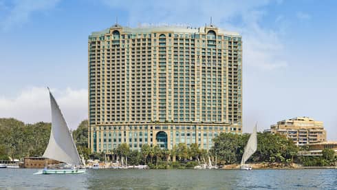 Exterior shot of a large hotel building on the water, sail boats can be seen in the foreground.