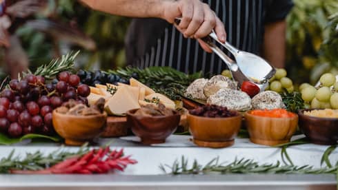 A chef's hand holds silver tongs over a display of grapes, dried fruits and cheese slices