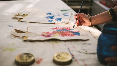 Close-up of a gold-bangled hand wielding a long brush to paint an intricate pattern on a cloth tote bag.