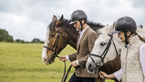 Man and woman wearing equestrian riding helmets lead horses through green field