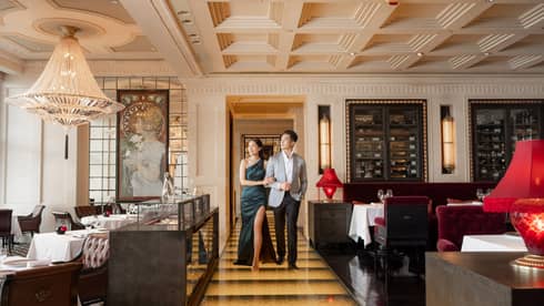 Elegant couple walking arm-in-arm in luxurious restaurant with chandeliers, red velvet seating and ornate decor.