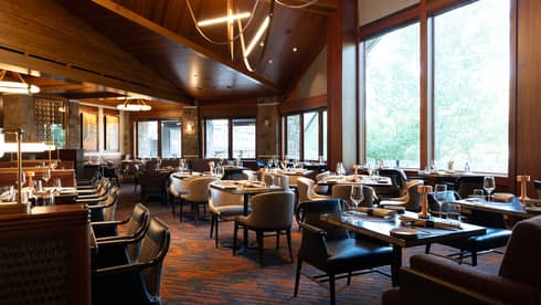 A restaurant with tables set with glasses and silverware, and windows along the sides of the room