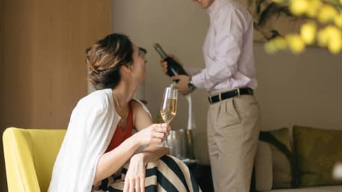 Person smiling and holding a glass of champagne while another person opens a bottle