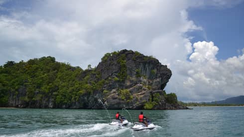 Two people wearing red life vests on jet skis ride on ocean under mountain