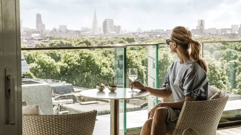 A guest takes in the city skyline while comfortably relaxing in a wicker chair and enjoying a drink on a balcony.