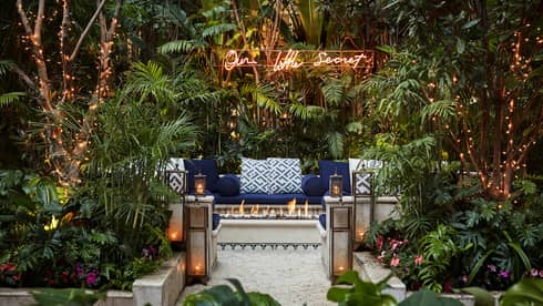 Our Little Secret sign over cushioned bench behind outdoor fireplace, lanterns, tropical garden