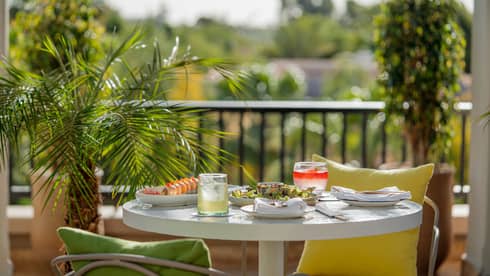 A sunlit outdoor dining table on a balcony topped with colourful dishes and cocktails