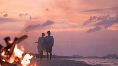Couple with Champagne glasses walks along sandy beach by bonfire at sunset