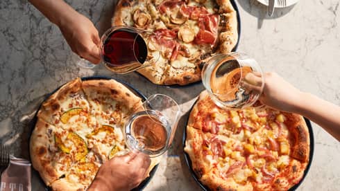 Three hands toast with glasses of wine above three whole pizzas at PIGNETO